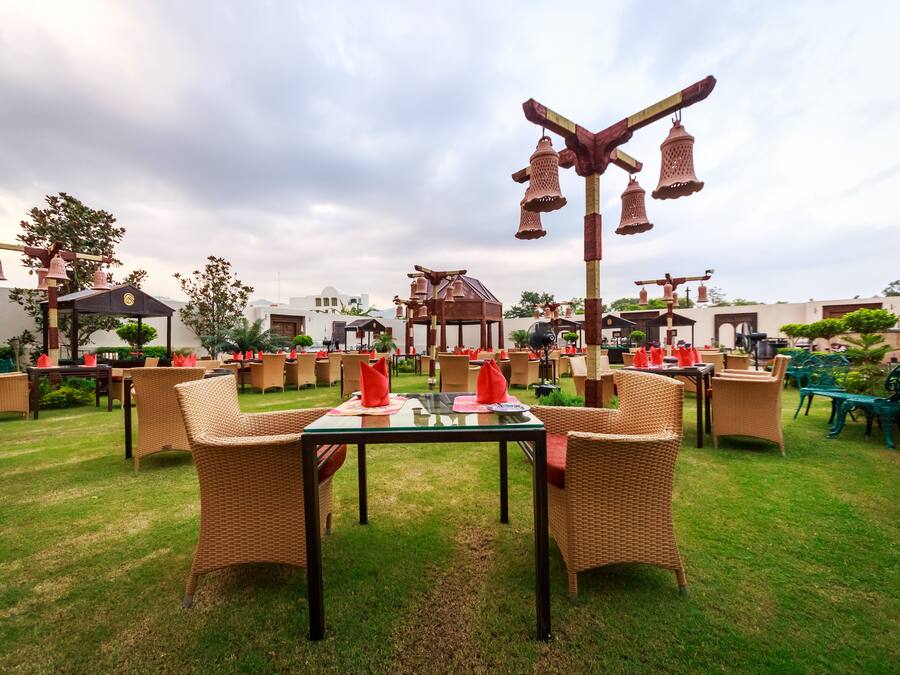 An outdoor dining area with wicker furniture and a central table, surrounded by greenery and a gazebo.