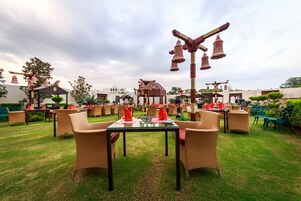An outdoor dining area with wicker furniture and a central table, surrounded by greenery and a gazebo.