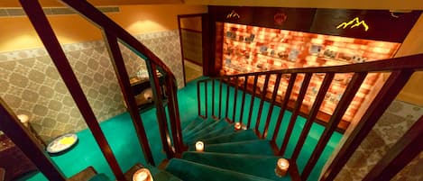 Una escalera con alfombra color turquesa y barandales de madera, que conduce a una habitación con paredes estampadas y una vitrina con diversos objetos.