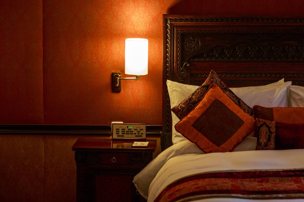 Una habitación de hotel con cabecera de madera, una mesita de noche con un reloj y una lámpara de pared.