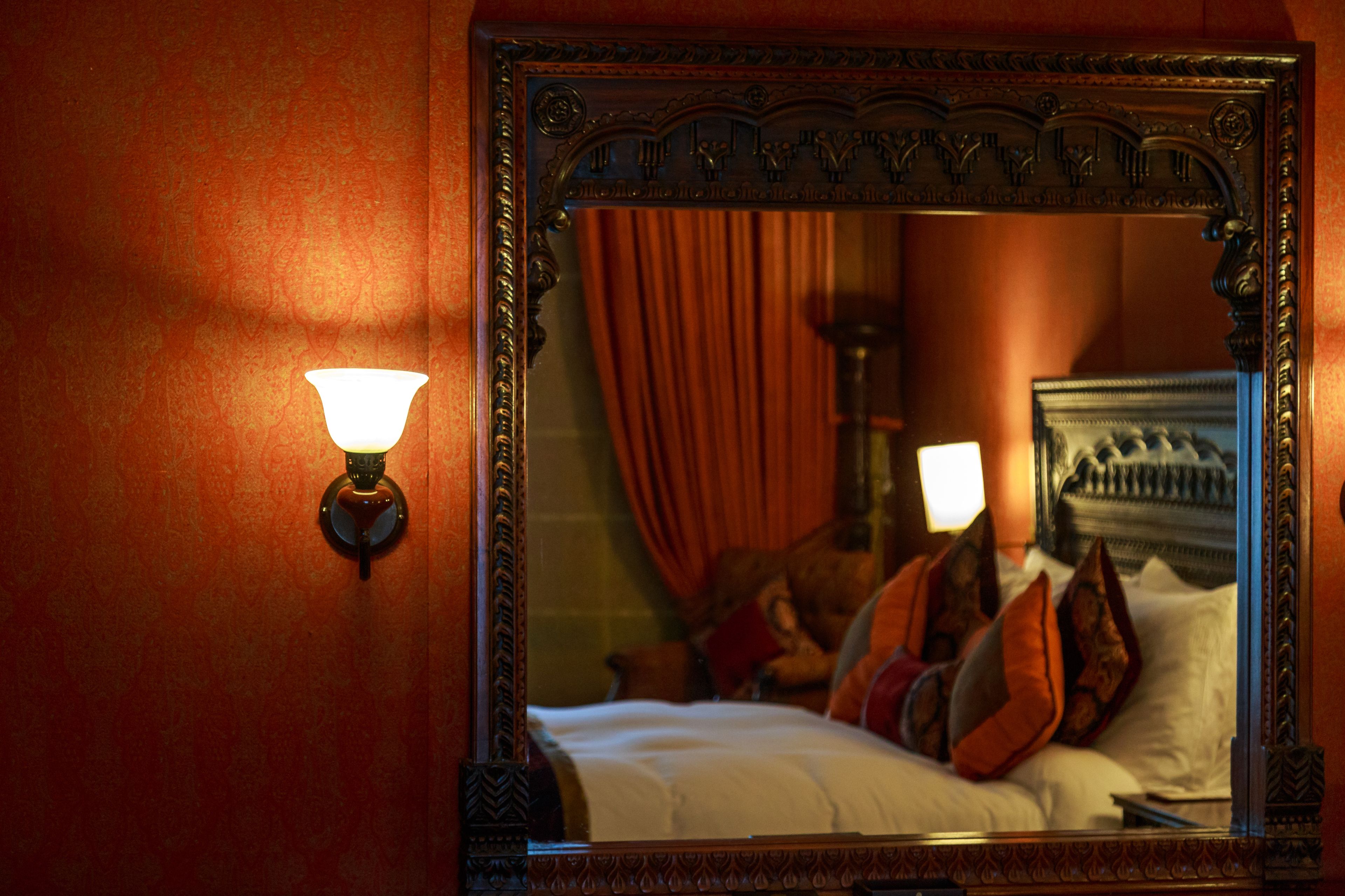 Uno specchio con una cornice decorata che riflette un letto con cuscini e una lampada.