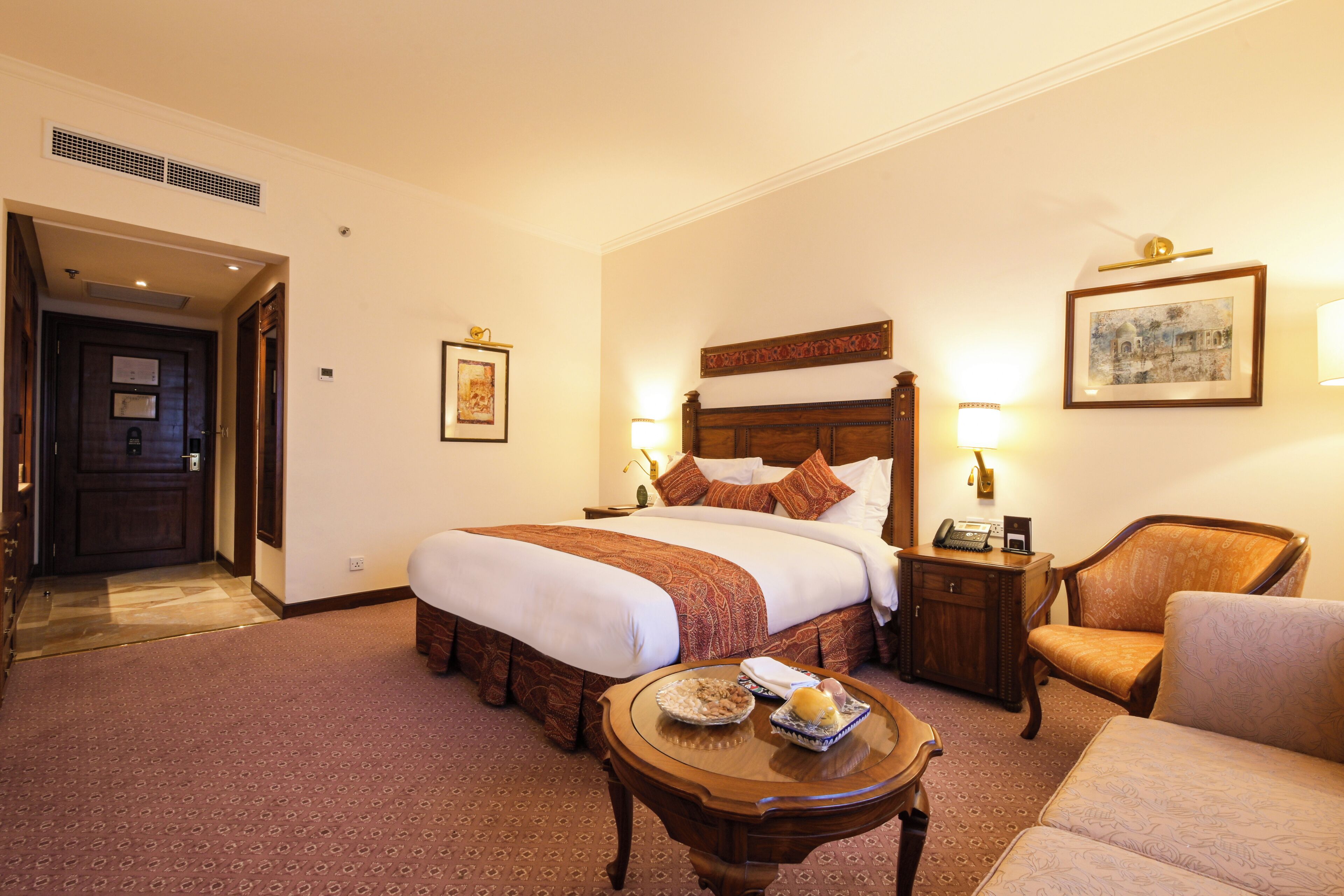 A hotel room with a large bed, bedside lamps, a wooden nightstand, a chair, and a small table with a bowl of cereal.