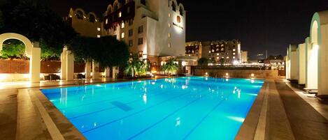 Una piscina con superficie de agua azul cristalina, rodeada de una zona de piscina bien iluminada y un edificio grande con varios pisos y balcones al fondo.