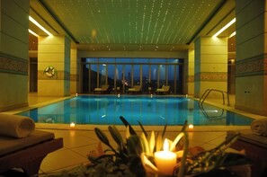 A well-lit indoor swimming pool with lounge chairs and a view of the cityscape at dusk. - Le Royal Hotel Amman (Amman)