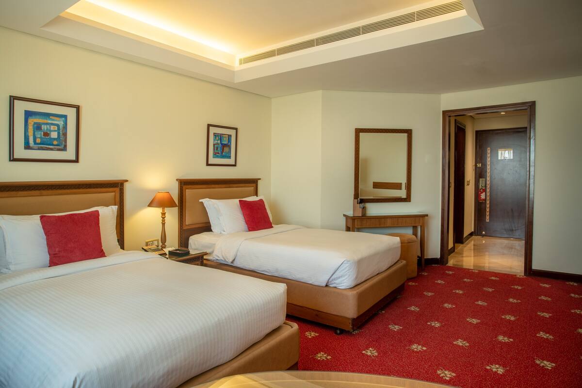 A hotel room with two beds, a red carpet, and a hallway leading to a door.