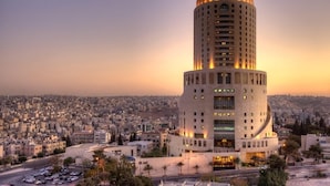 A tall, cylindrical hotel with a curved lower section, surrounded by a cityscape. - Le Royal Hotel Amman (Amman)