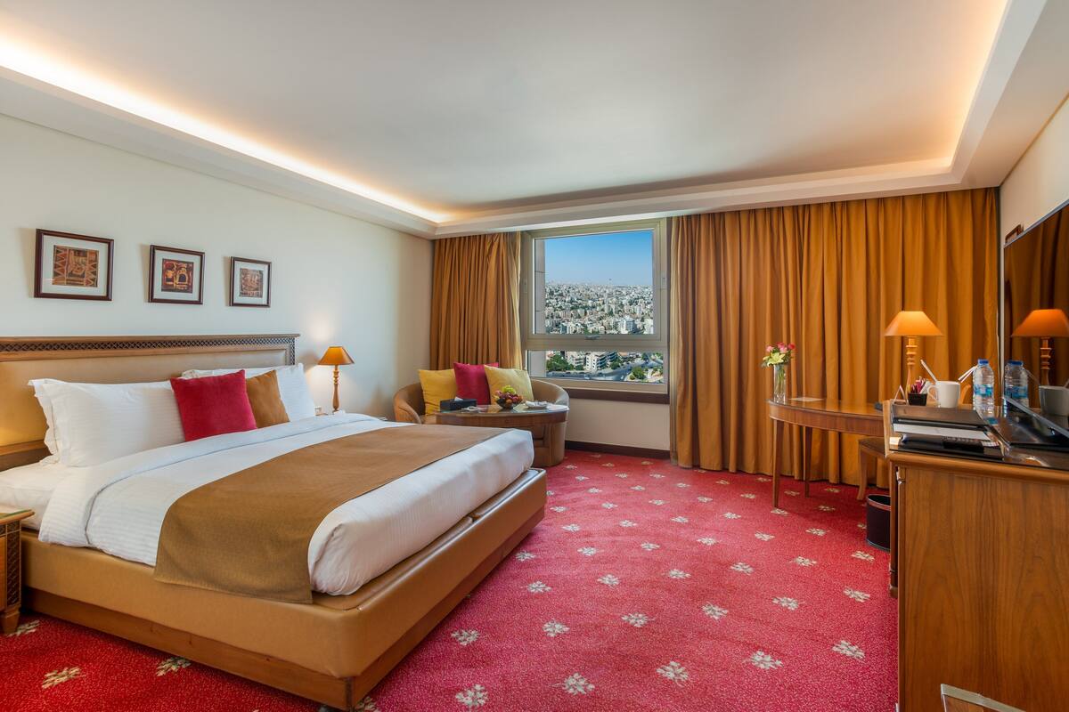A hotel room with a large bed, a desk, a chair, a window with curtains, and framed pictures on the wall.