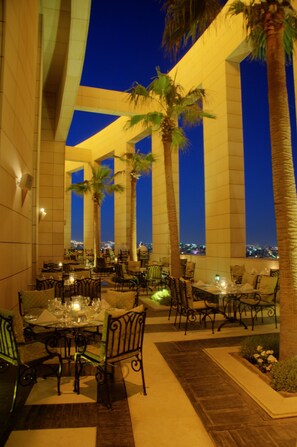 An outdoor dining area with tables and chairs, palm trees, and a city view at dusk. - Le Royal Hotel Amman (Amman)
