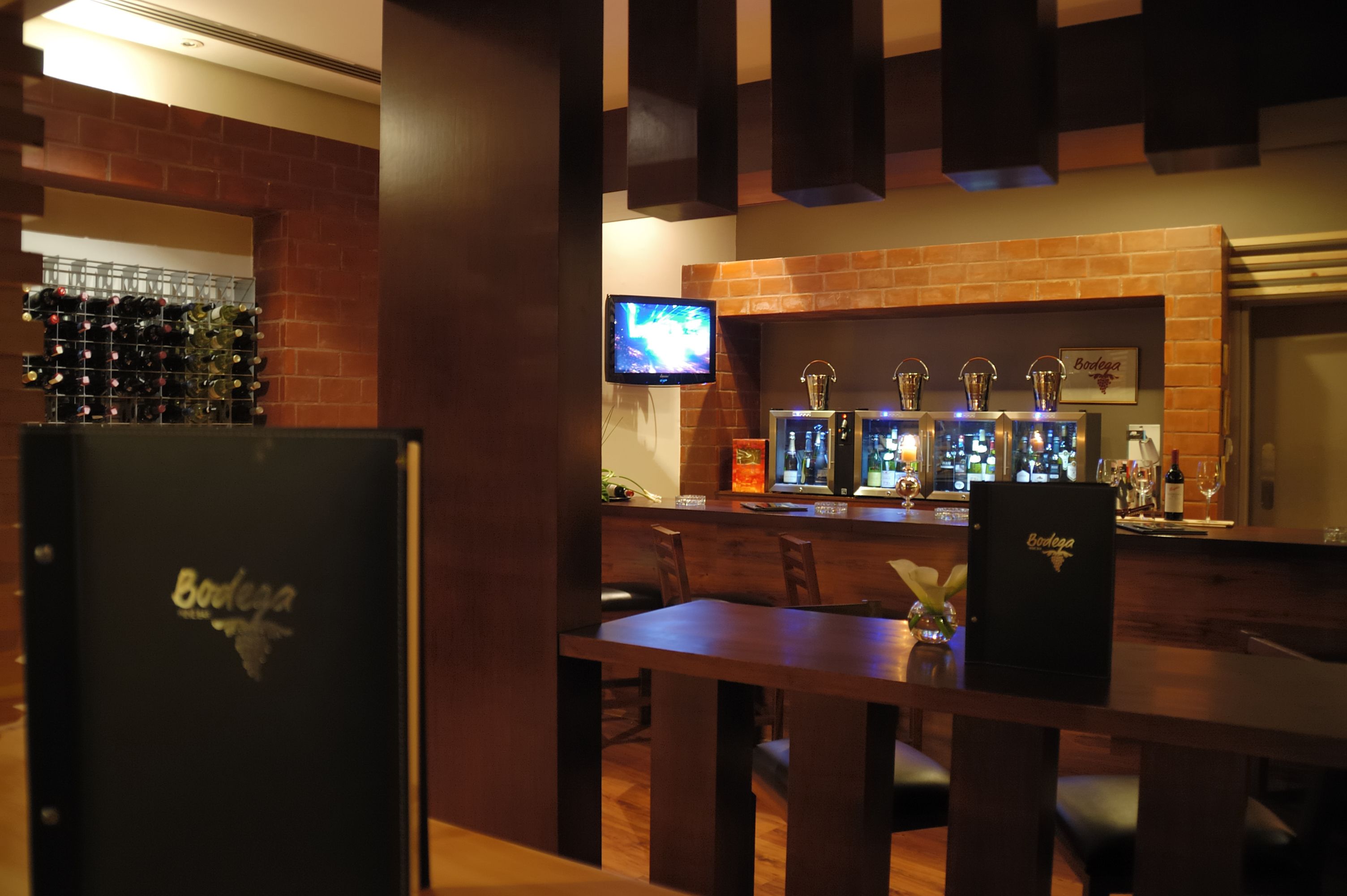 A bar area with a brick wall, a television mounted above the bar, and a wine rack filled with bottles.