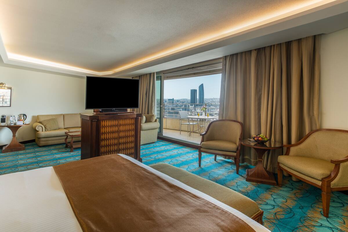 A hotel room with a large bed, a television, a sofa, a chair, a small table with a fruit bowl, and a view of the cityscape through large windows.