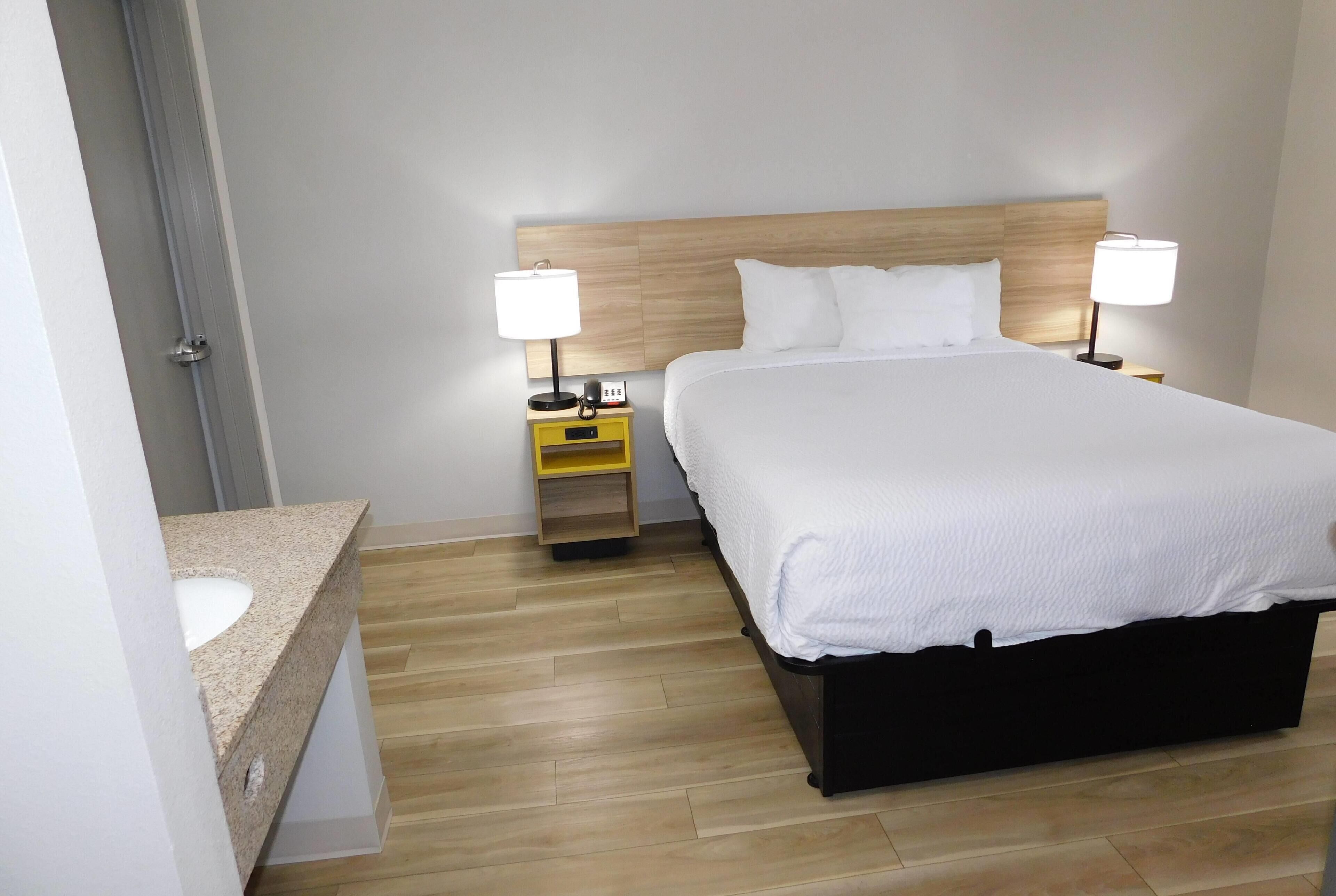A hotel room with a bed, bedside tables, a nightstand, a bathroom with a sink, and wooden flooring.