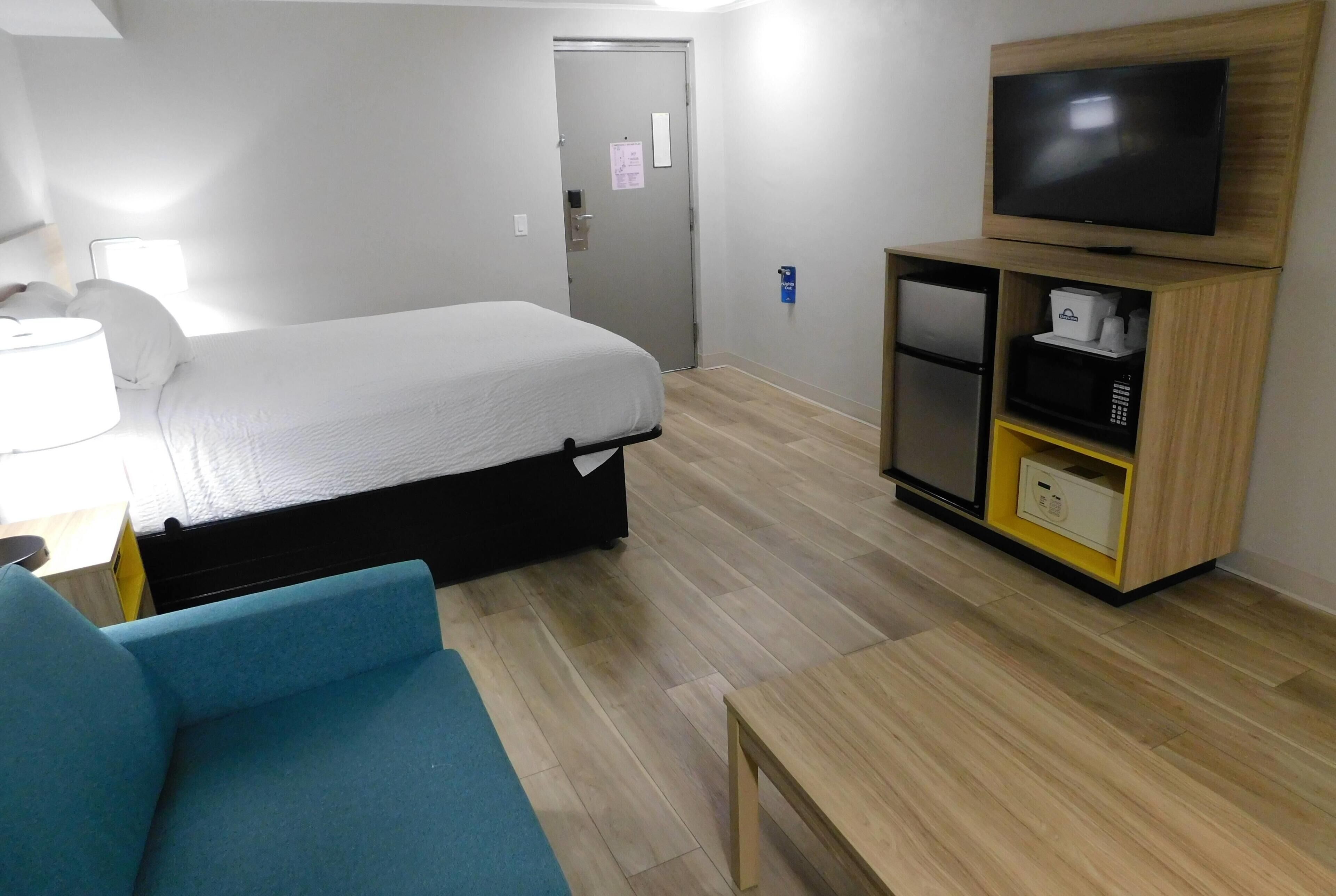 A hotel room with a bed, a TV, a desk with a lamp, a blue chair, and a wooden cabinet with a microwave.