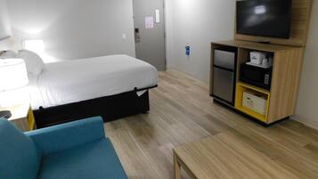 A hotel room with a bed, a TV, a desk with a lamp, a blue chair, and a wooden cabinet with a microwave.