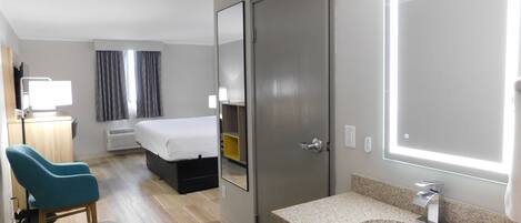 A hotel room with a bed, a desk with a computer, a chair, a bathroom with a sink and mirror, and a door.