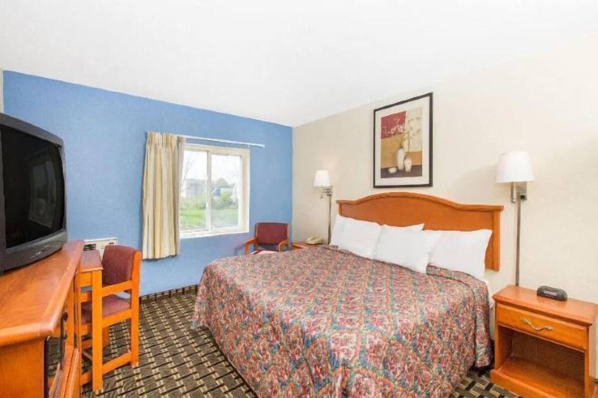 A hotel room with a bed, a desk with a television, a chair, a window with curtains, and a wall-mounted picture.