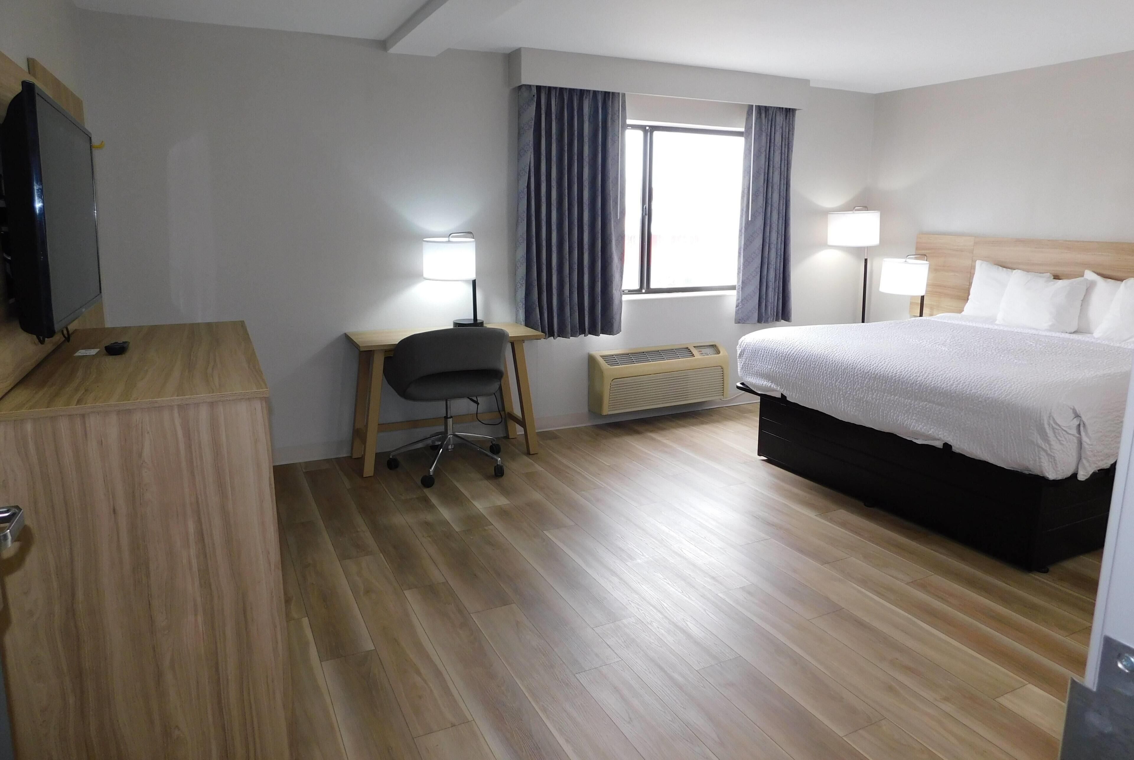 A hotel room with a wooden desk, a chair, a bed with white bedding, a TV, and a window with curtains.