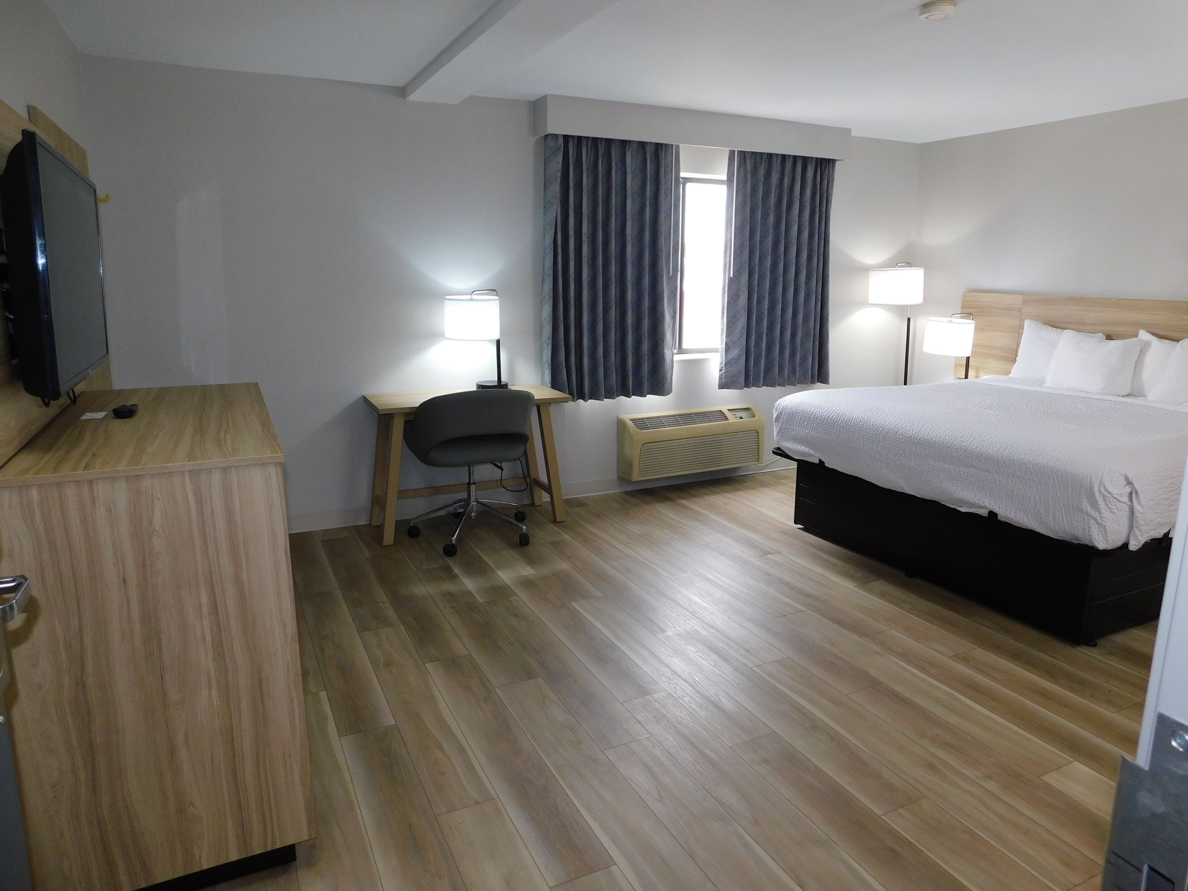 A hotel room with a bed, desk, chair, television, two bedside lamps, and a window with curtains.
