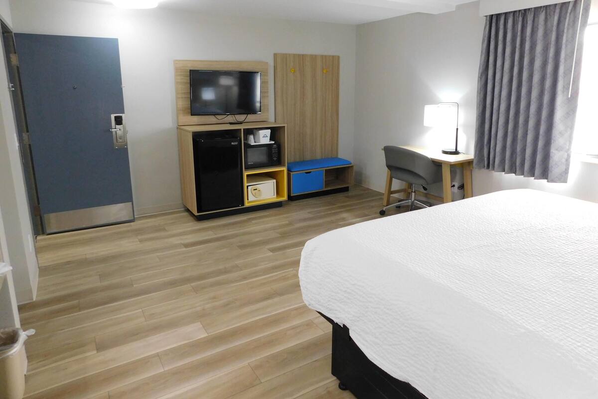 A hotel room with a bed, a TV, a desk, a chair, a wardrobe, and a small storage unit.