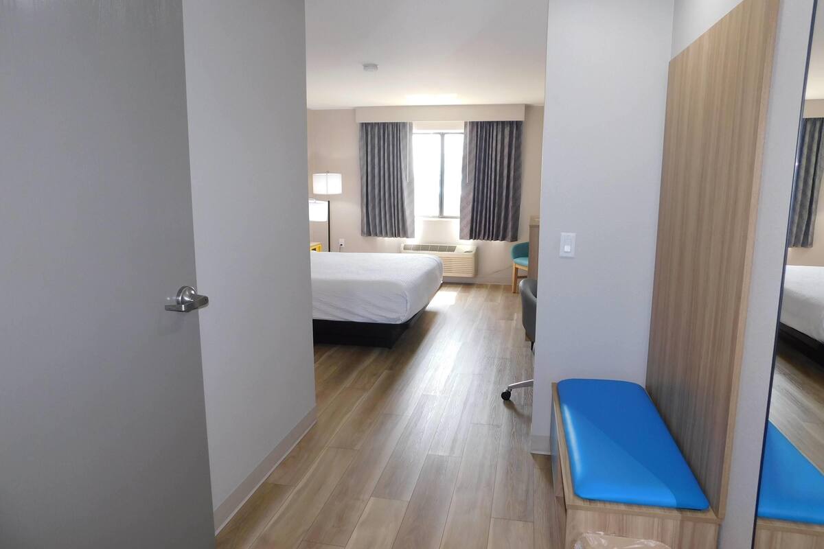A hotel room with a bed, a desk, a chair, a bench, a window with curtains, and a mirror.