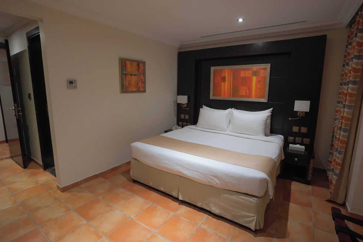 A hotel room with a large bed, two bedside tables with lamps, a painting on the wall, and a door to the left.