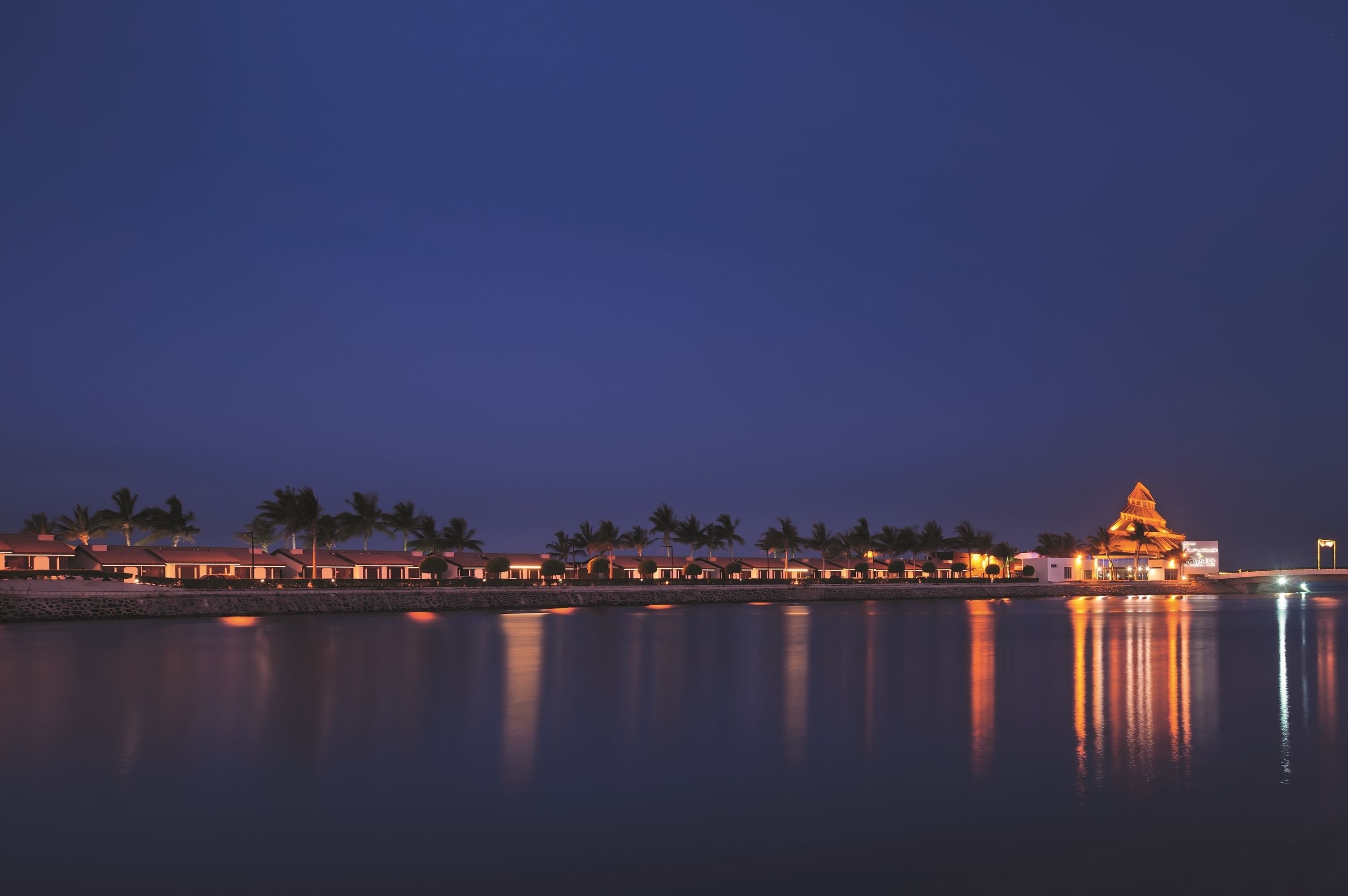 Un hotel frente al mar con un edificio destacado de techo dorado, palmeras y una superficie acuática tranquila y reflectante.