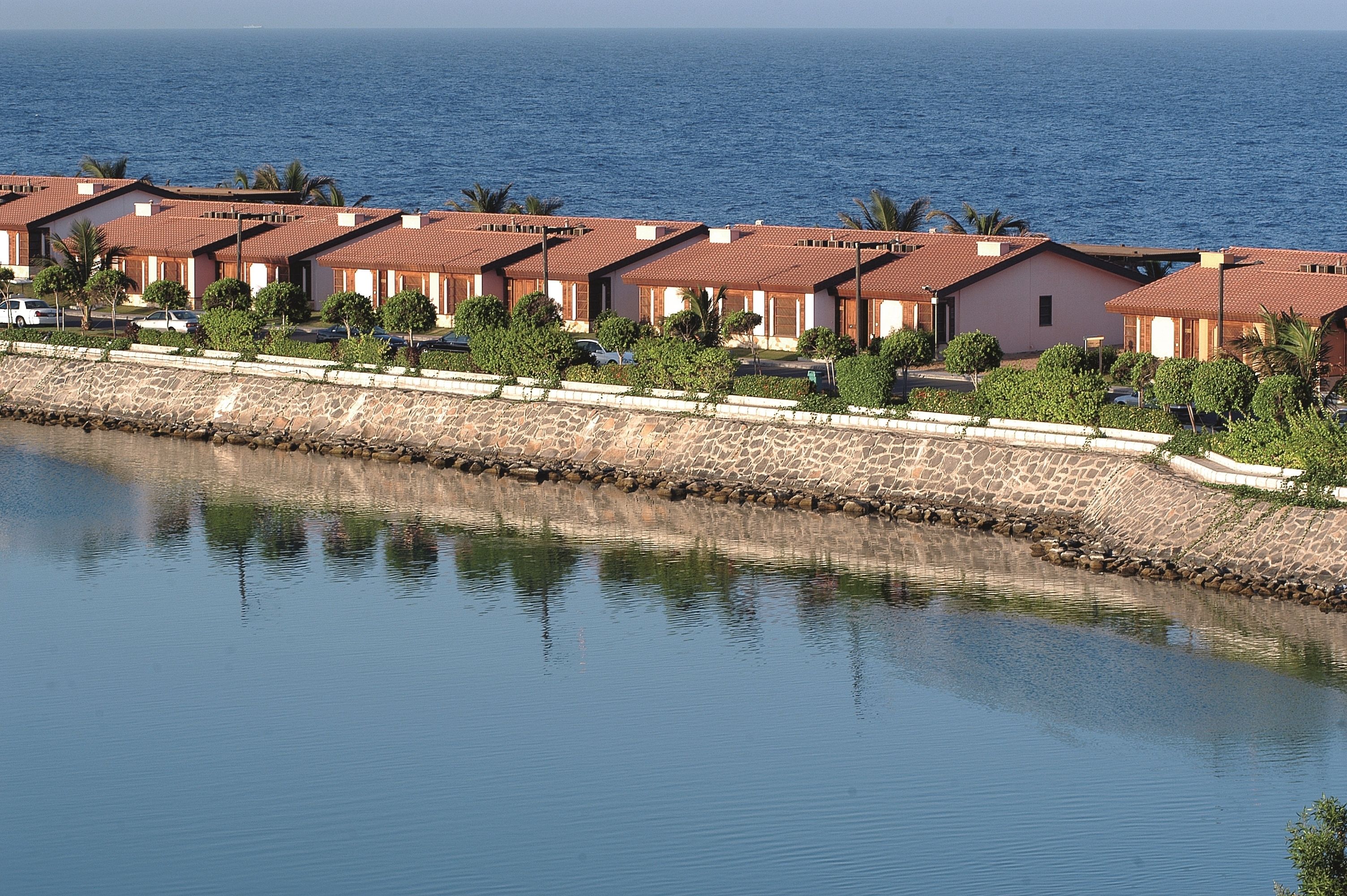 Un hôtel situé sur la côte, avec des bâtiments aux toits de tuiles rouges, un mur de pierre et une mer calme en arrière-plan.