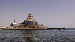 Un edifici costaner amb una cúpula central, una construcció moderna a l'esquerra i un litoral rocos.