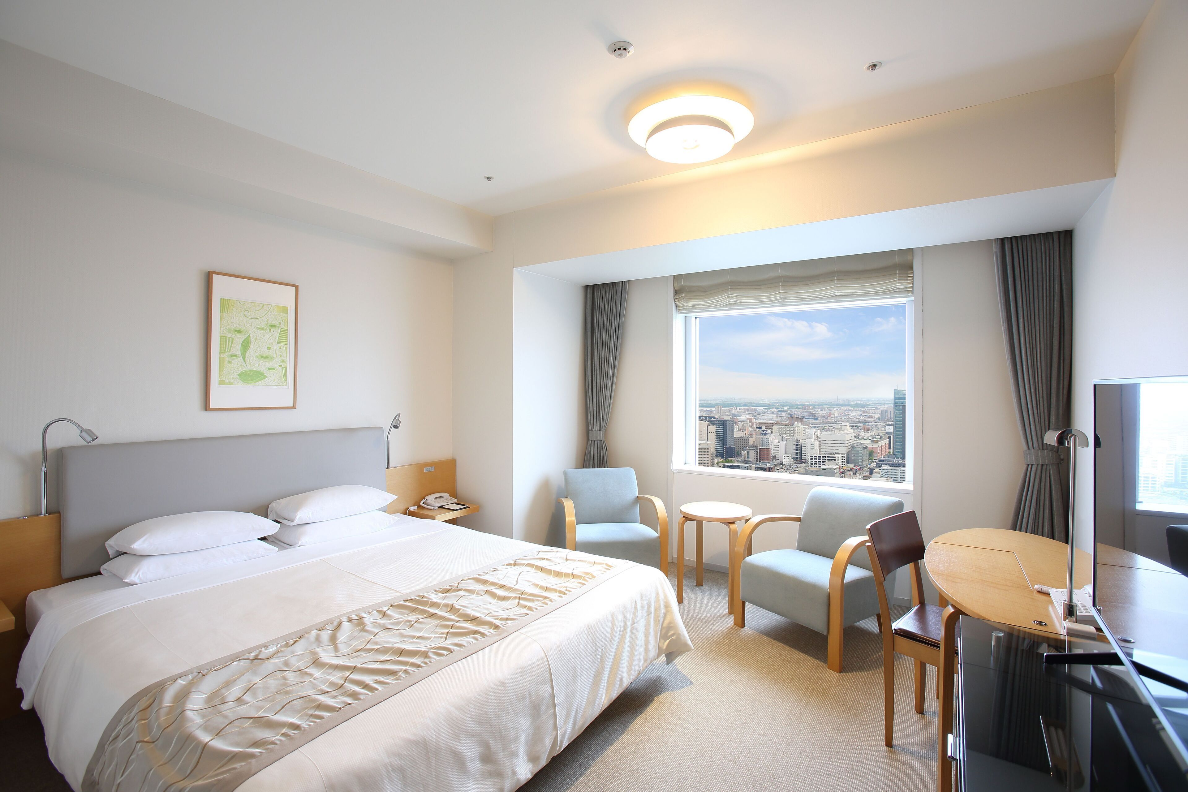 Habitación de hotel con cama, dos sillas, una mesita, escritorio, lámpara, ventana con cortinas y vistas a la ciudad.
