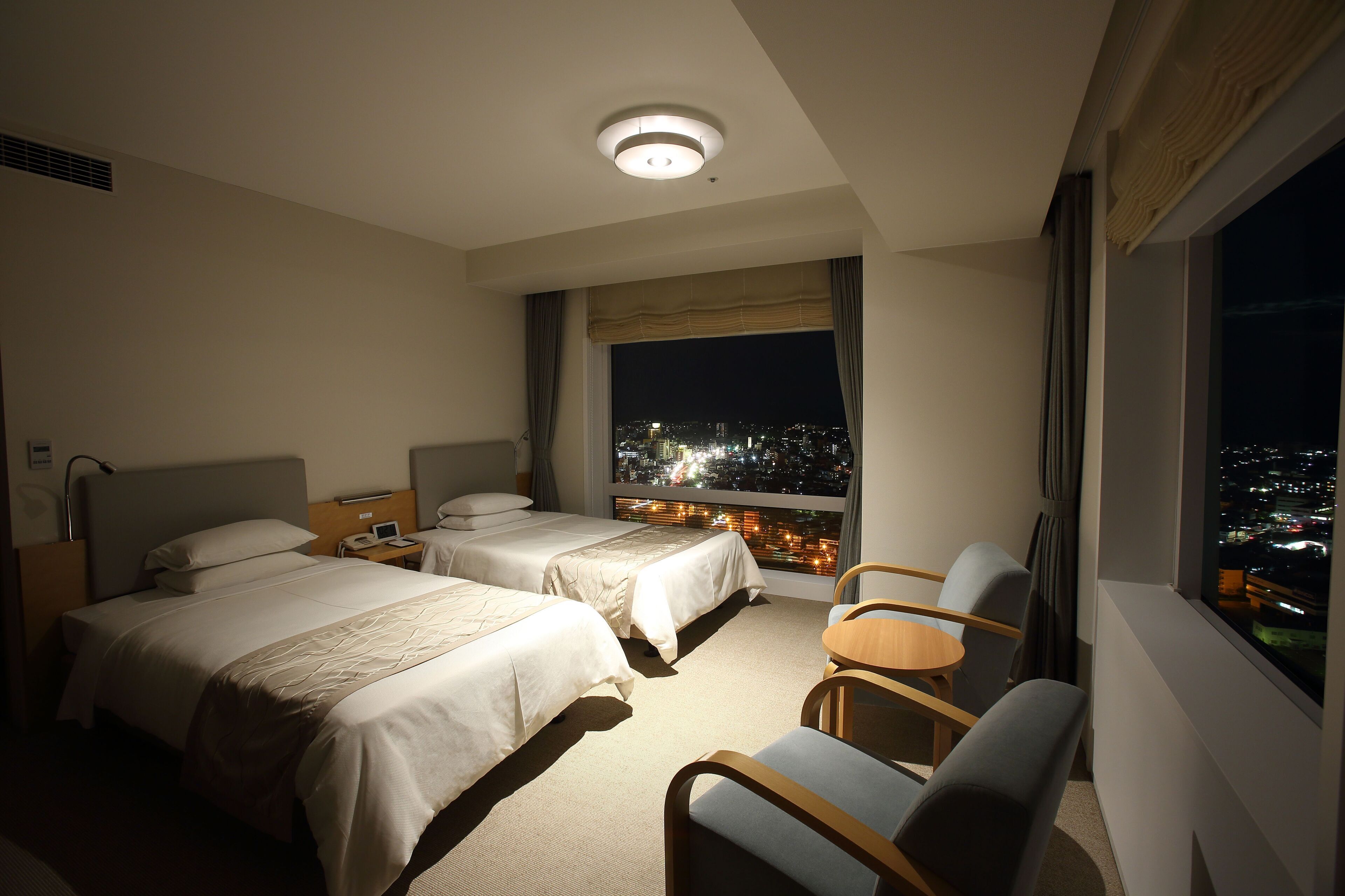 A hotel room with two beds, a small table, and a chair. There is a window with a city view at night.