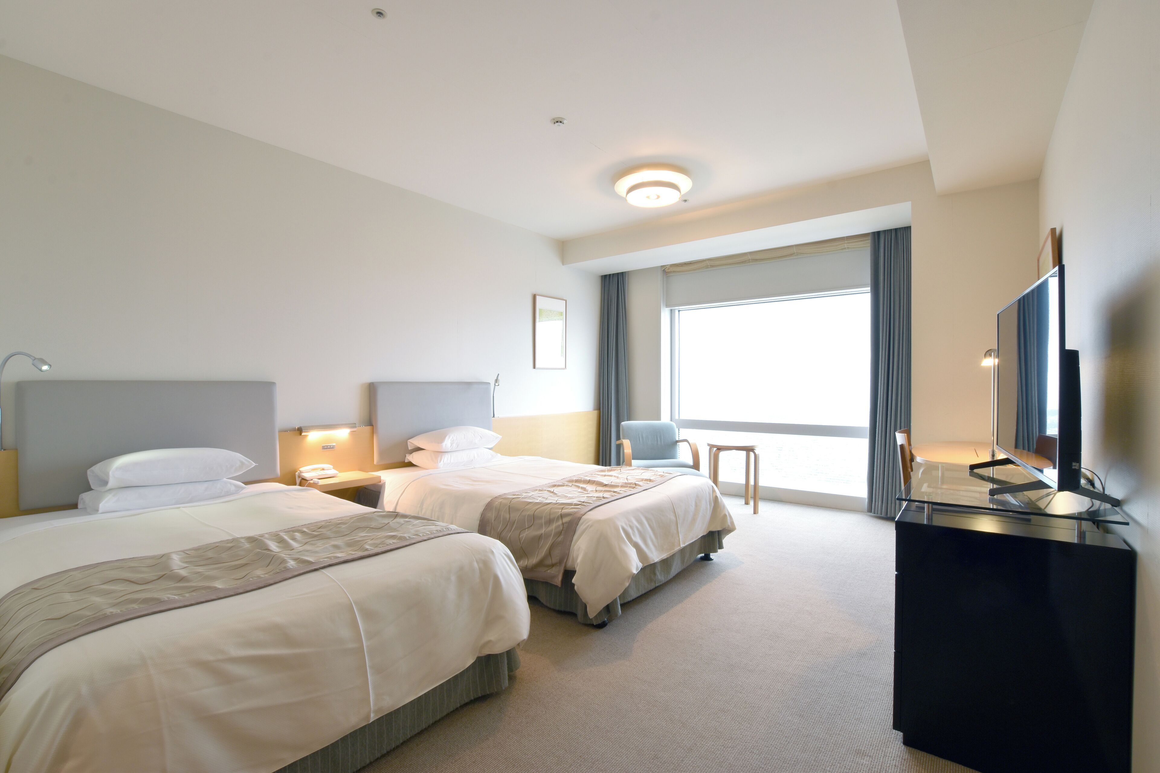 A hotel room with two beds, a desk, a chair, a TV, and a large window with curtains.