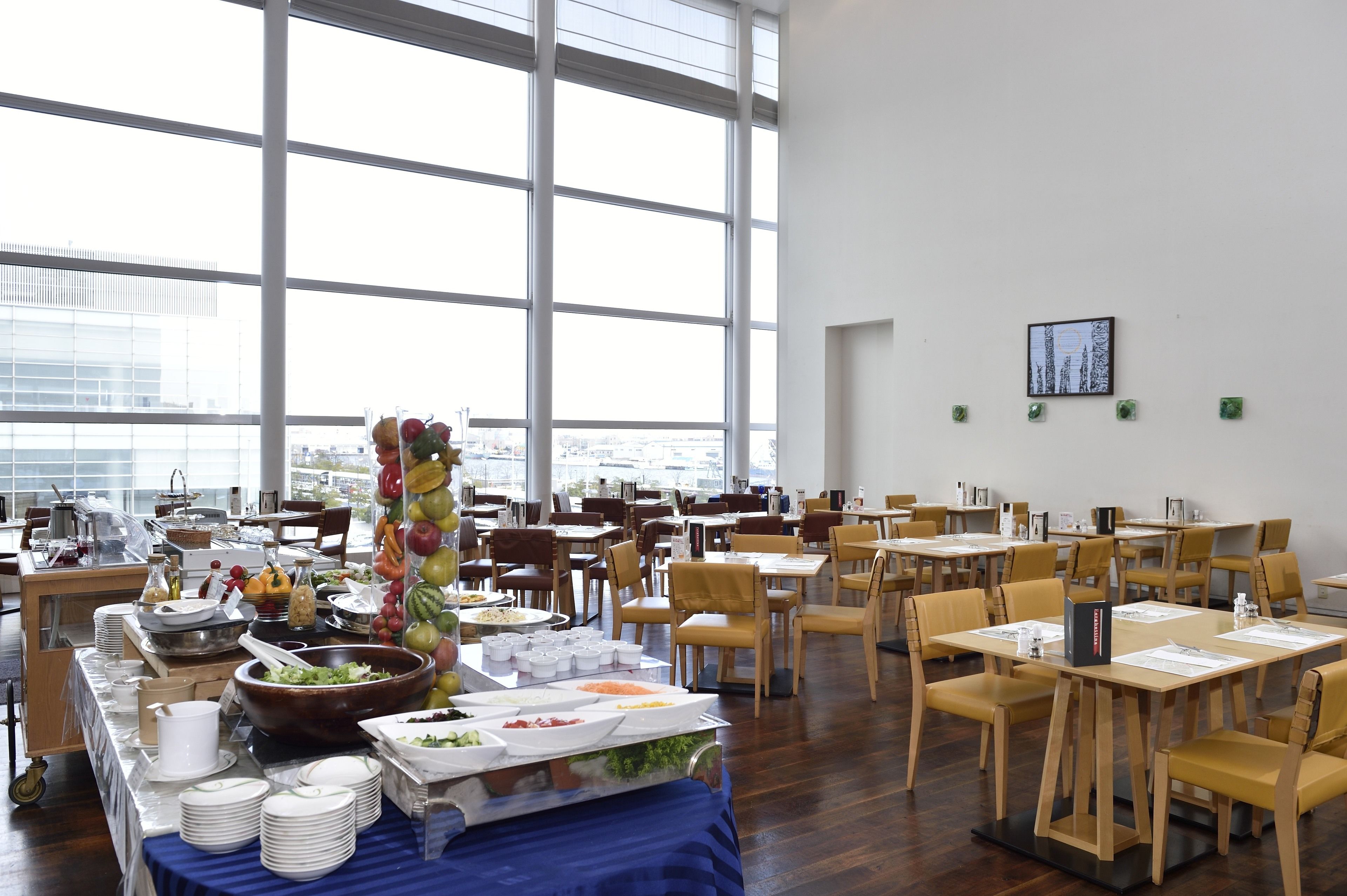 A dining area with tables and chairs, a buffet with various food items, and large windows offering a view of the city.