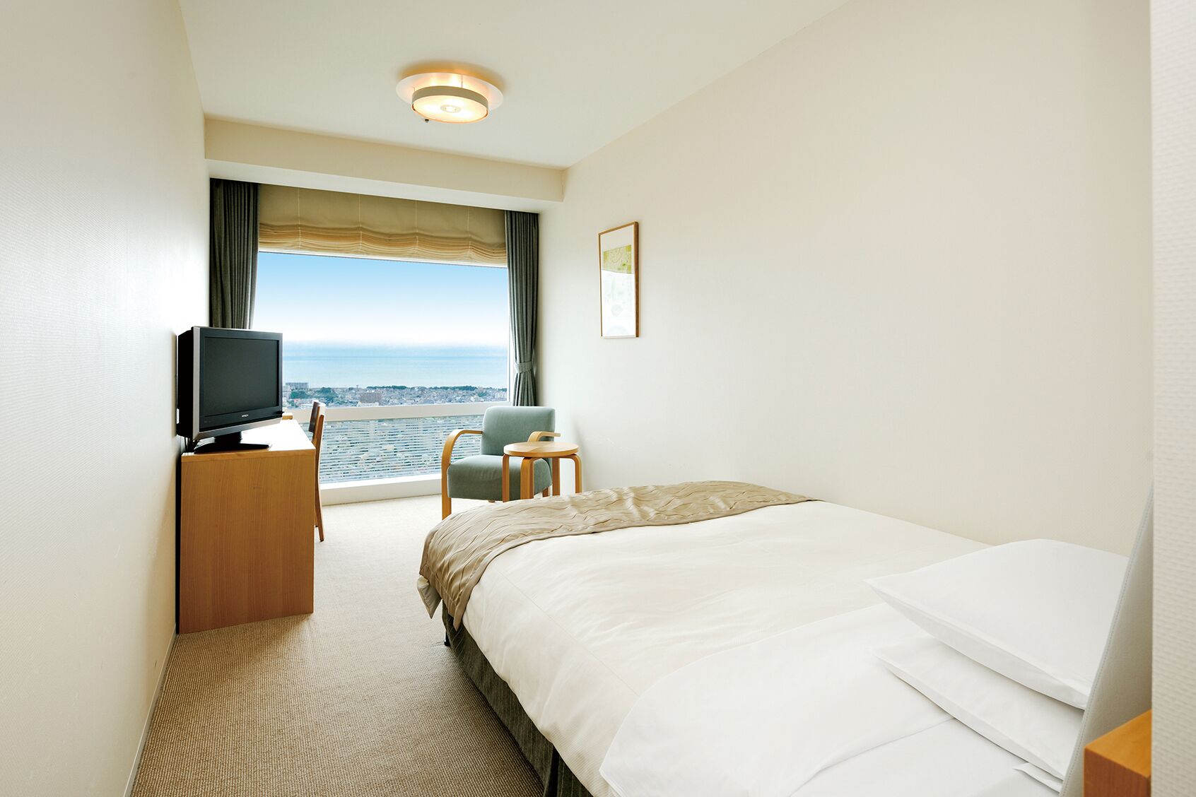 A hotel room with a bed, a desk with a television, a chair, a small table, and a window with a view of the city and ocean.