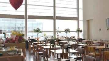 A spacious dining area with multiple tables and chairs, large windows, and potted plants.