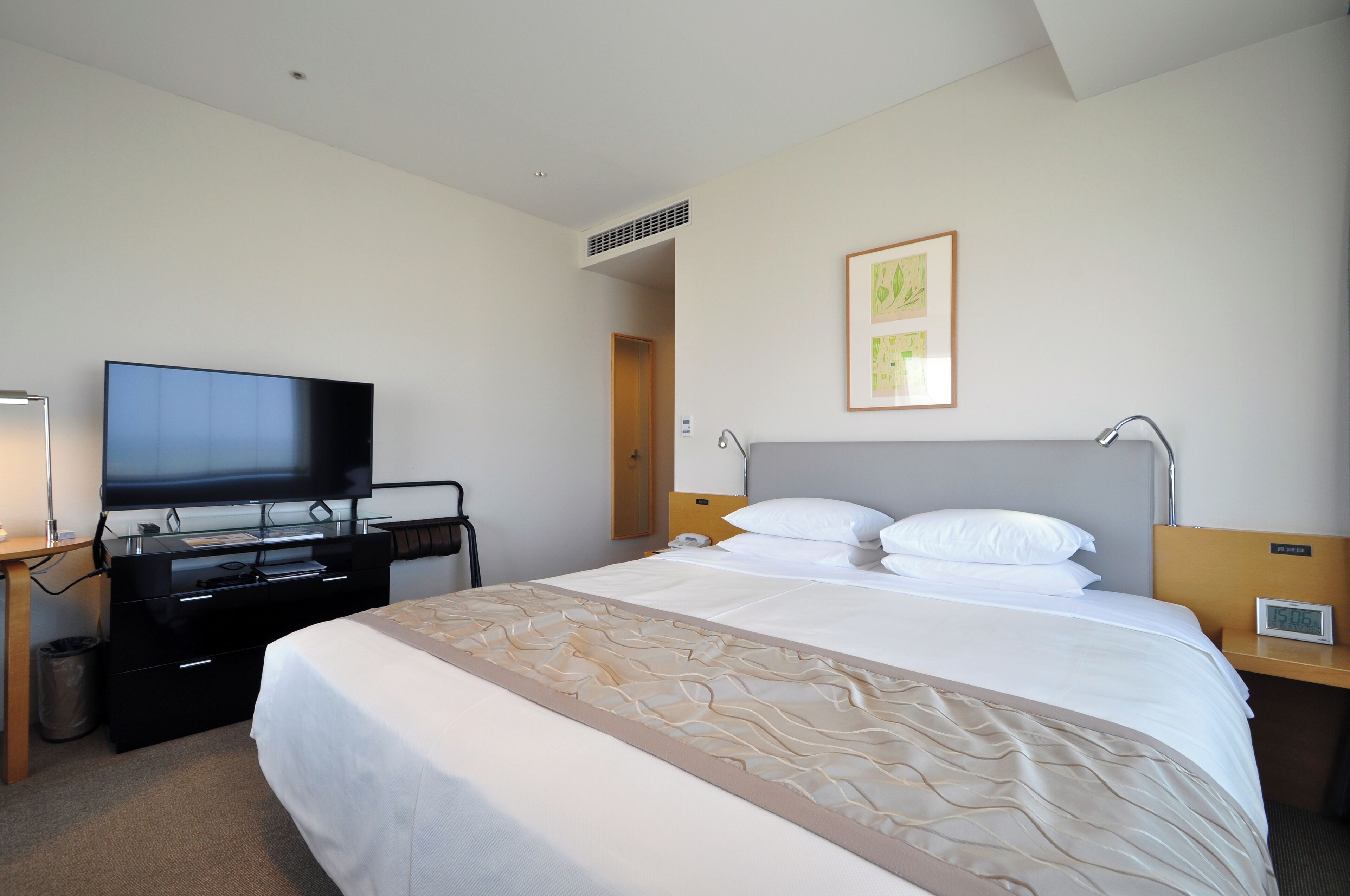 A hotel room with a bed, a TV, a desk with a lamp, and a wall-mounted artwork.