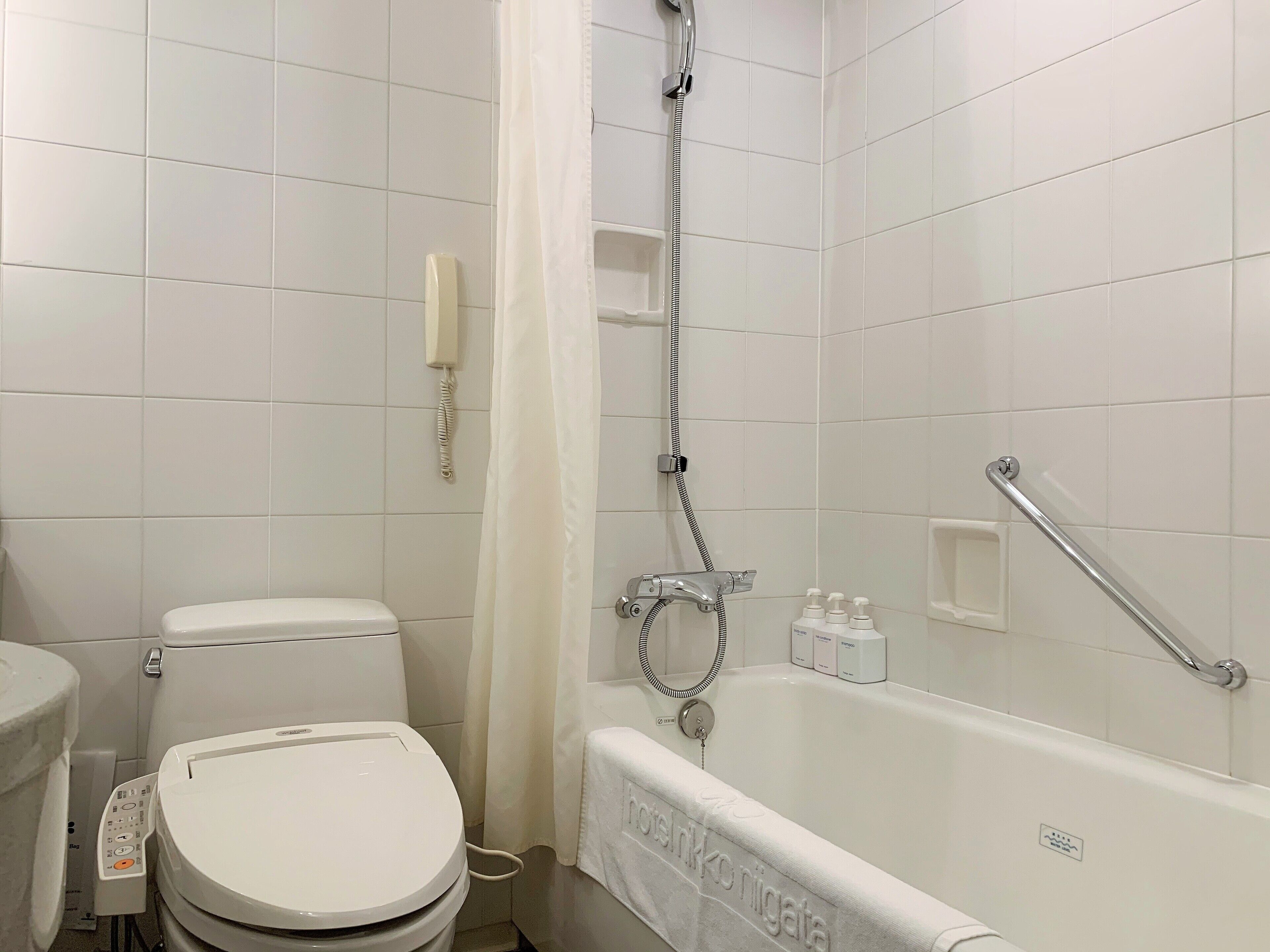 A bathroom with a toilet, a shower with a handheld showerhead, a towel rack, and a soap dispenser.