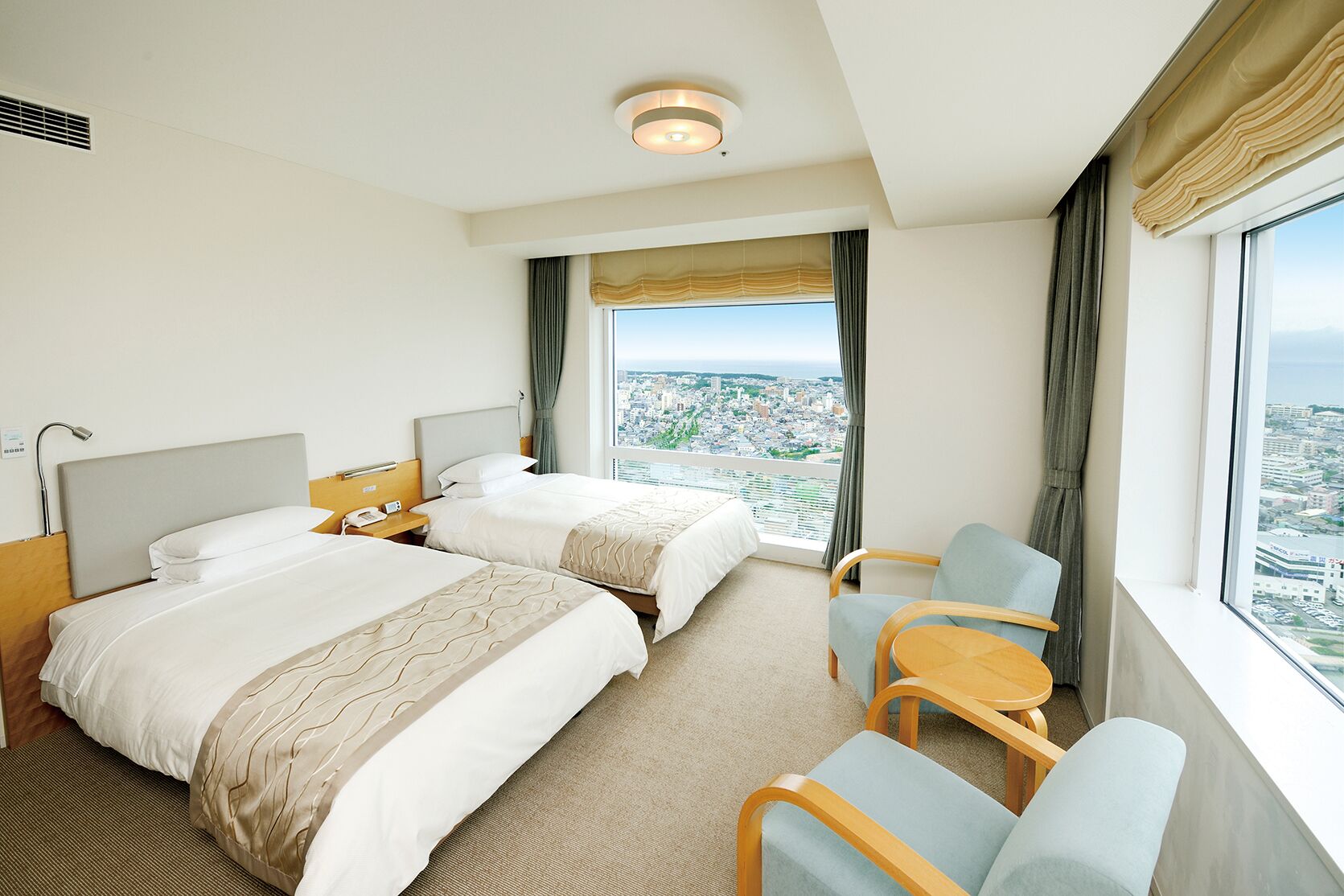 Habitación de hotel con dos camas, zona de estar con una mesa y sillas, vista a un paisaje urbano y un ventanal grande con cortinas.