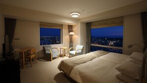A hotel room with a large bed, two armchairs, a small table, and a desk with a lamp. There are two windows with city views. - Hotel Nikko Niigata (Niigata)