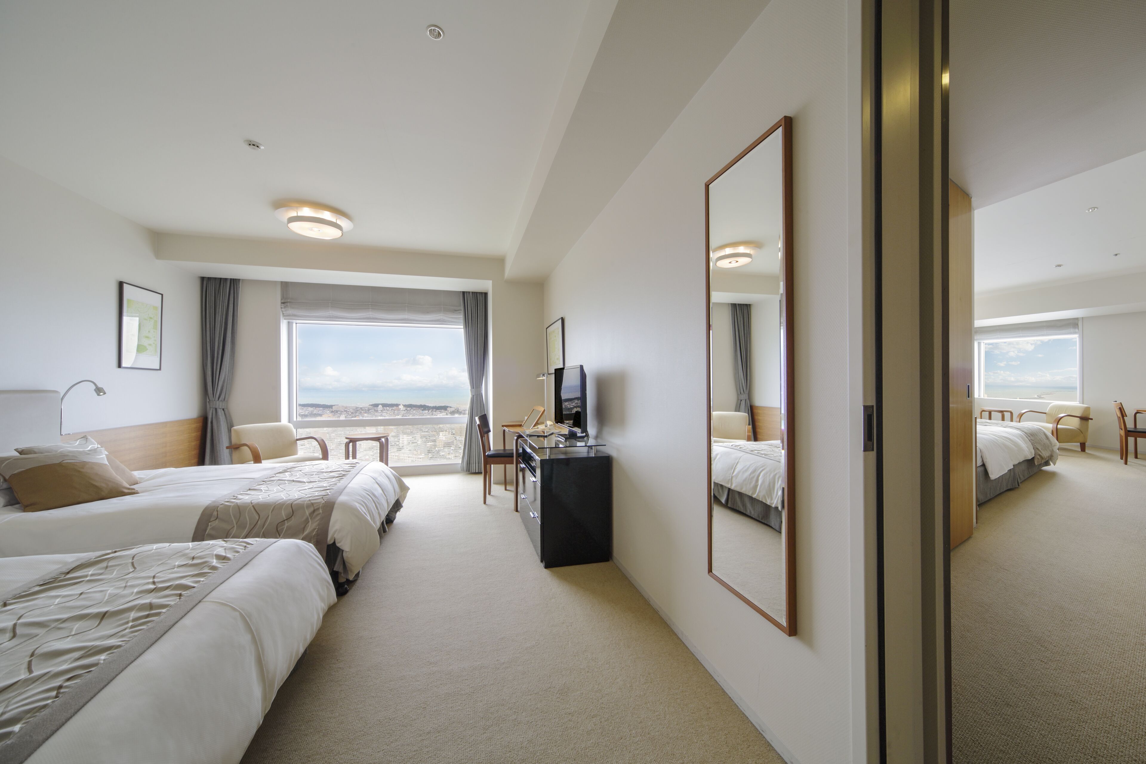A hotel room with two beds, a desk, a TV, and a large window offering a view.