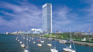 Una marina con varios veleros amarrados, un edificio moderno y alto al fondo, y un cielo azul despejado.