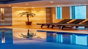 Un área de piscina interior moderna con una palmera en maceta, sillas de descanso y una piscina cristalina que refleja el techo.