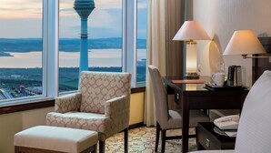Habitación de hotel con un ventanal que ofrece vista a una torre y una extensión de agua. Hay dos sillas, una mesa con una lámpara y un teléfono.