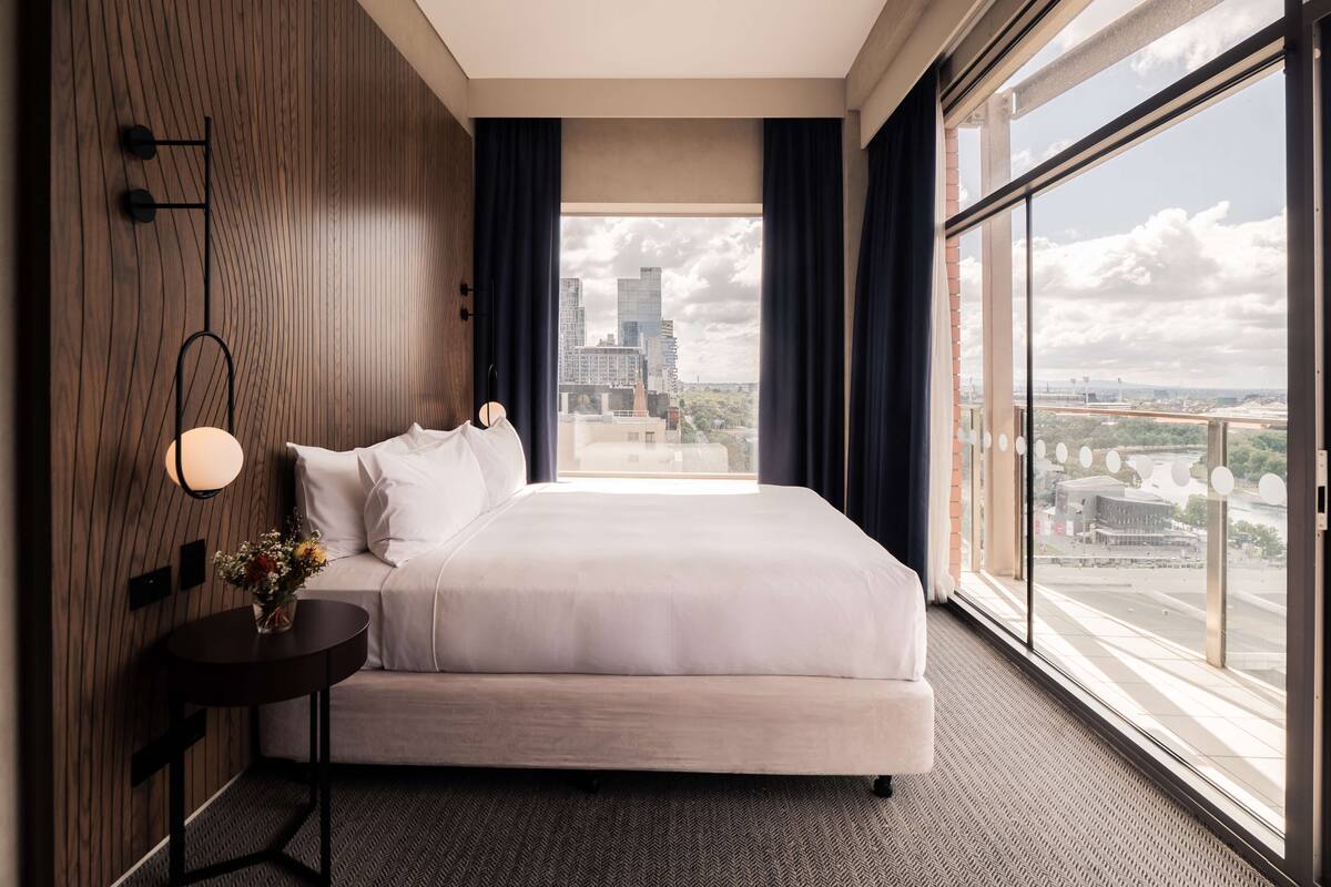 A modern hotel room with a large bed, a round side table with a vase of flowers, a wall-mounted lamp, and a large window offering a city view.