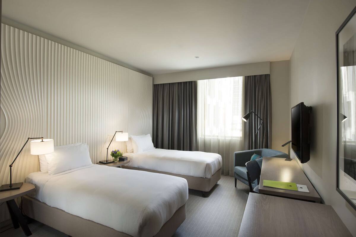 A hotel room with two beds, a desk, a chair, a TV, and a window with curtains.