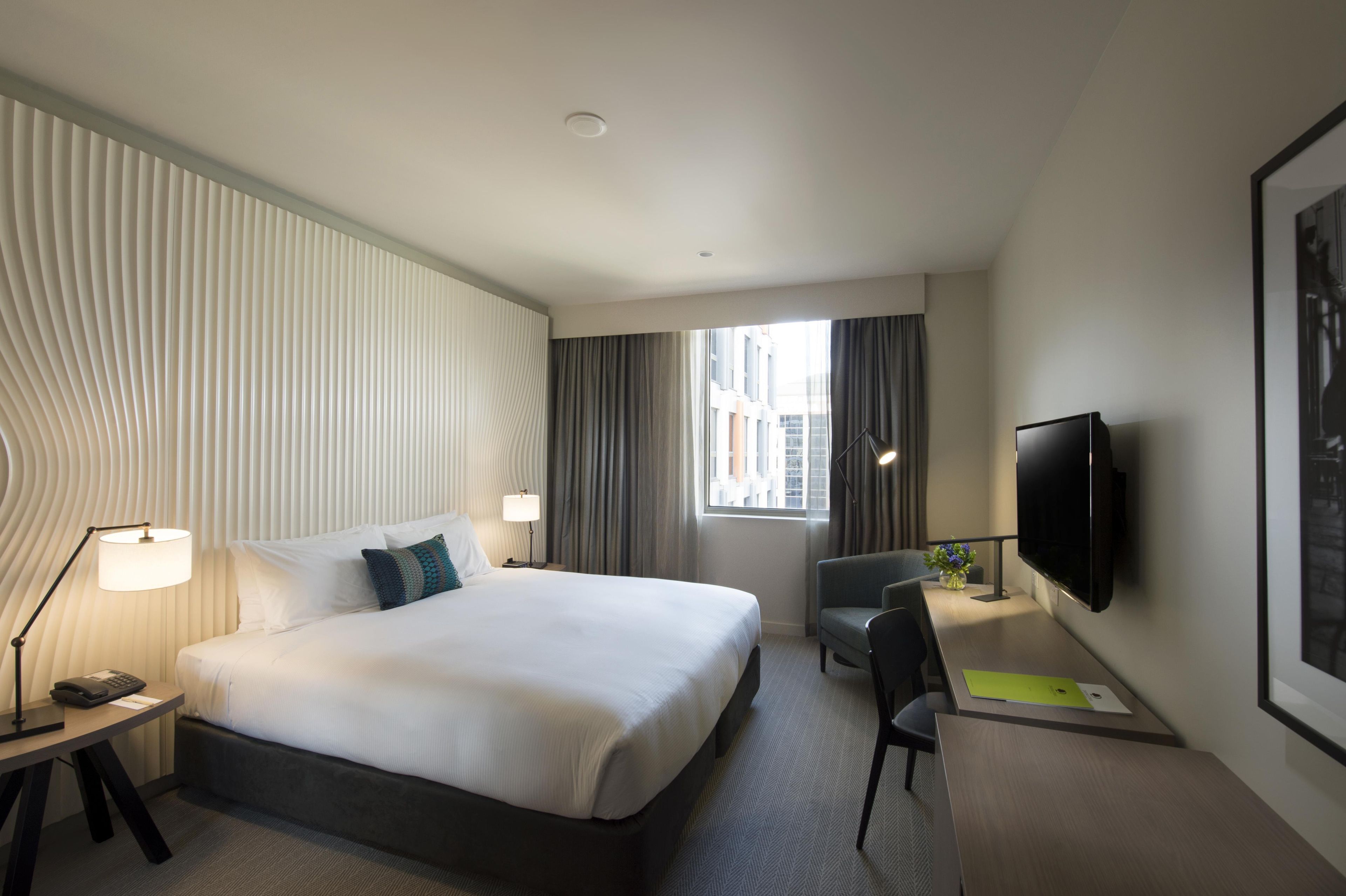 A hotel room with a large bed, a desk with a chair, a flat-screen TV, and a window with curtains.