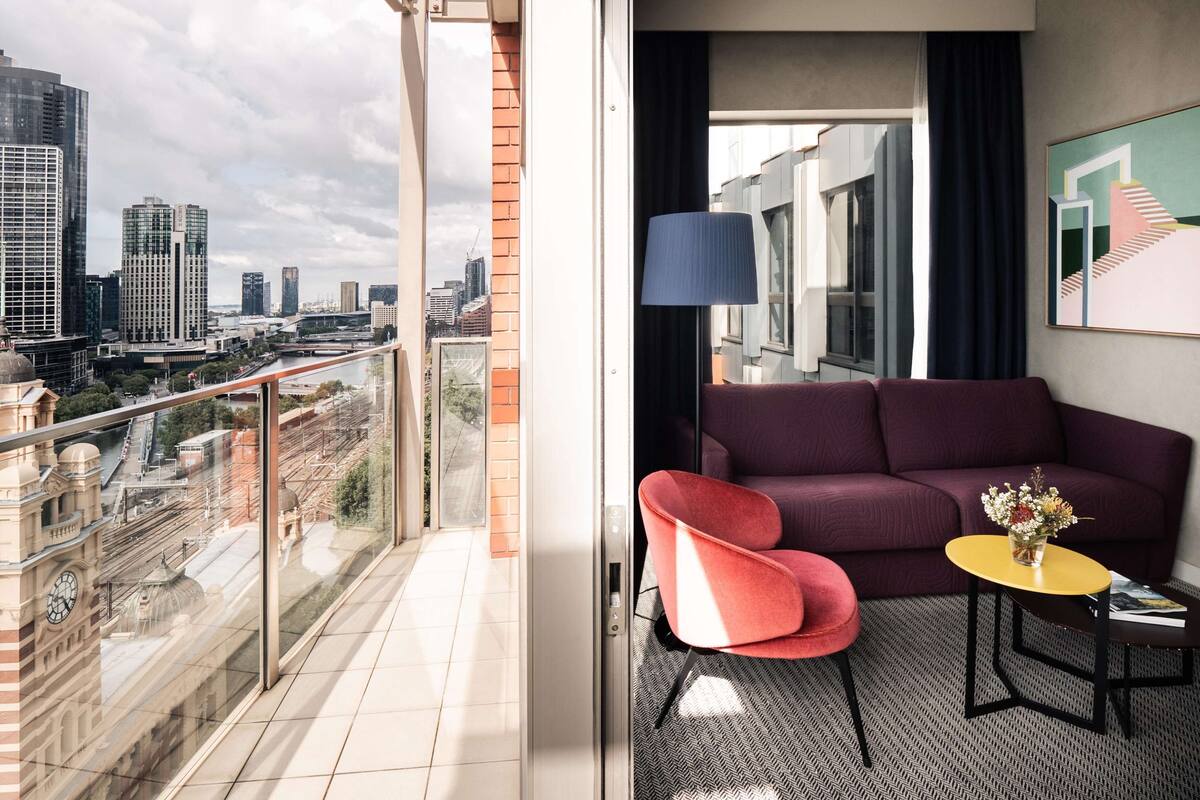 A modern hotel room with a balcony offering a city view, a comfortable seating area with a sofa and armchair, and a small table with a vase of flowers.
