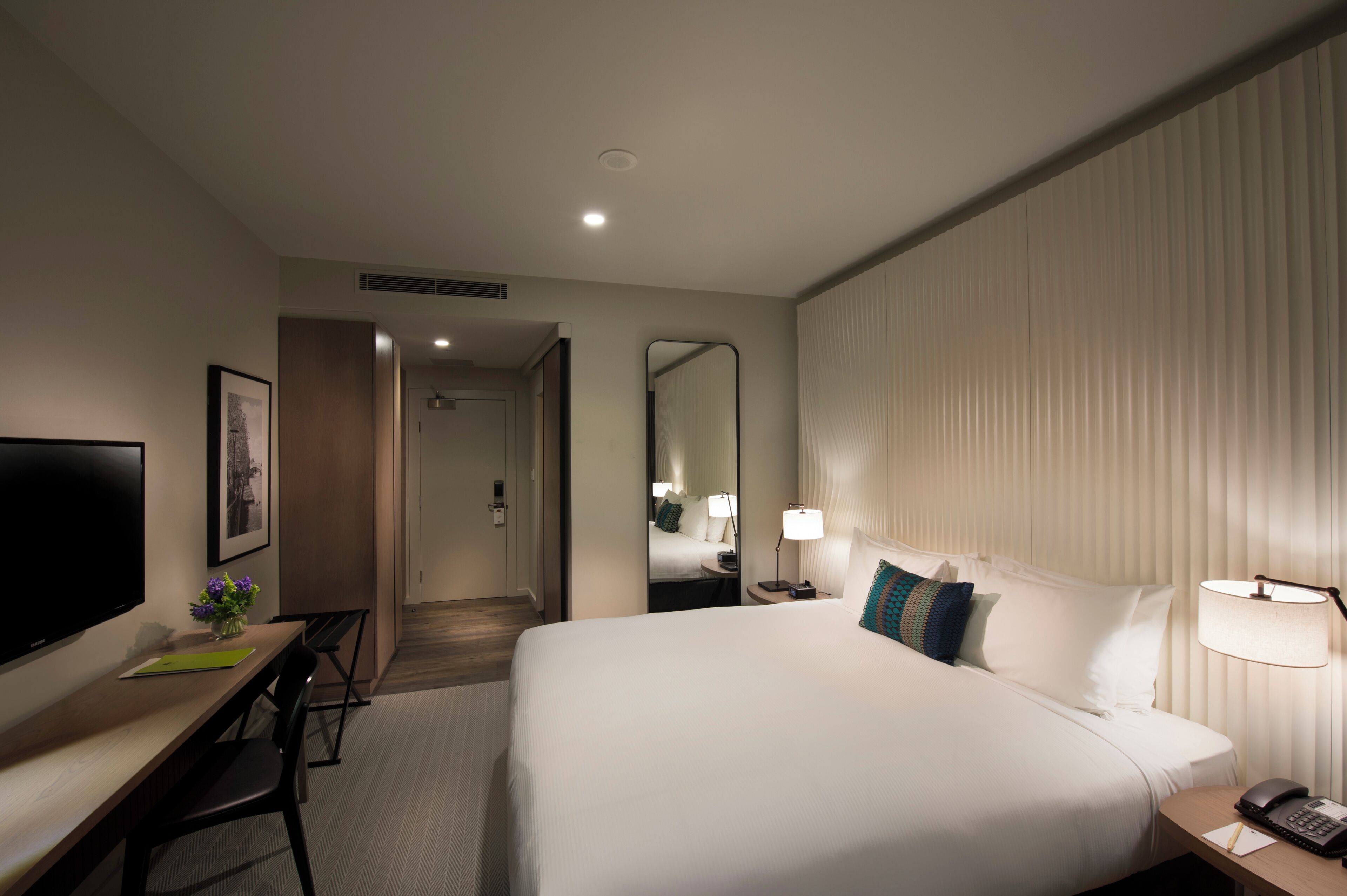 A hotel room with a large bed, a desk with a flat-screen TV, a chair, a lamp, and a telephone.