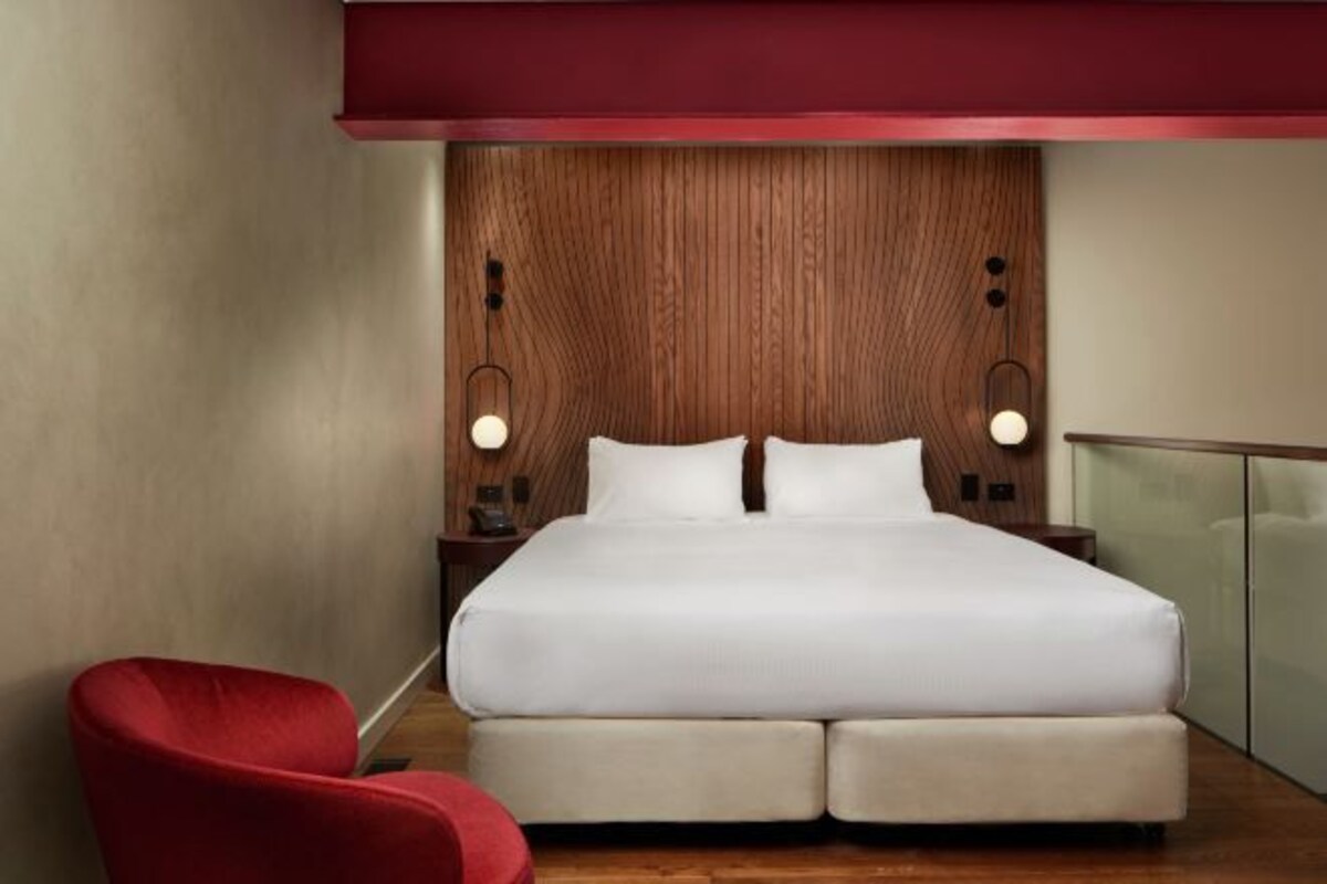 Una camera d'hotel moderna con un letto grande, una testiera in legno, due comodini con lampade e una sedia rossa.