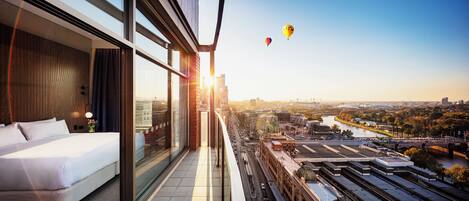 Une chambre d’hôtel située dans un immeuble de grande hauteur, dotée d’un grand lit, d’un mur en verre et offrant une vue sur le paysage urbain et la rivière.