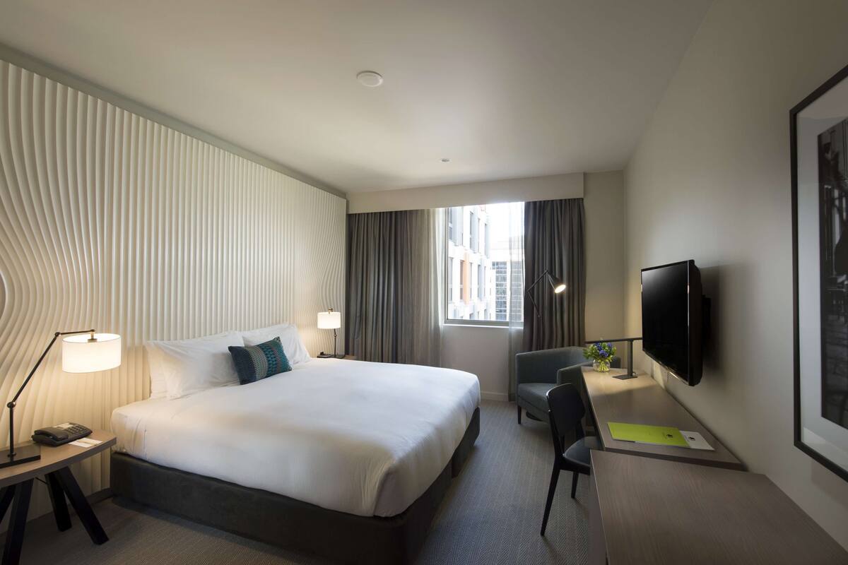 A hotel room with a large bed, a desk with a chair, a flat-screen TV, and a window with curtains.