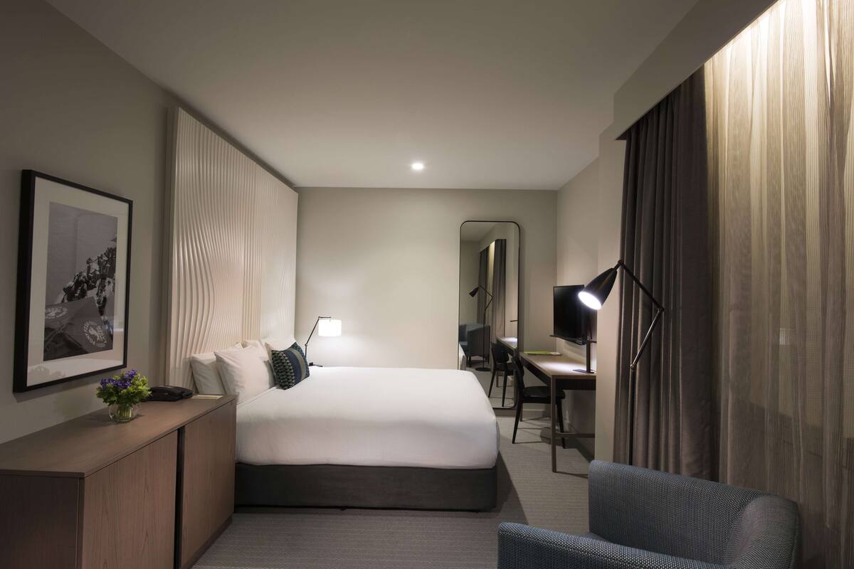 A modern hotel room with a large bed, a desk, a chair, a desk lamp, and a framed picture on the wall.